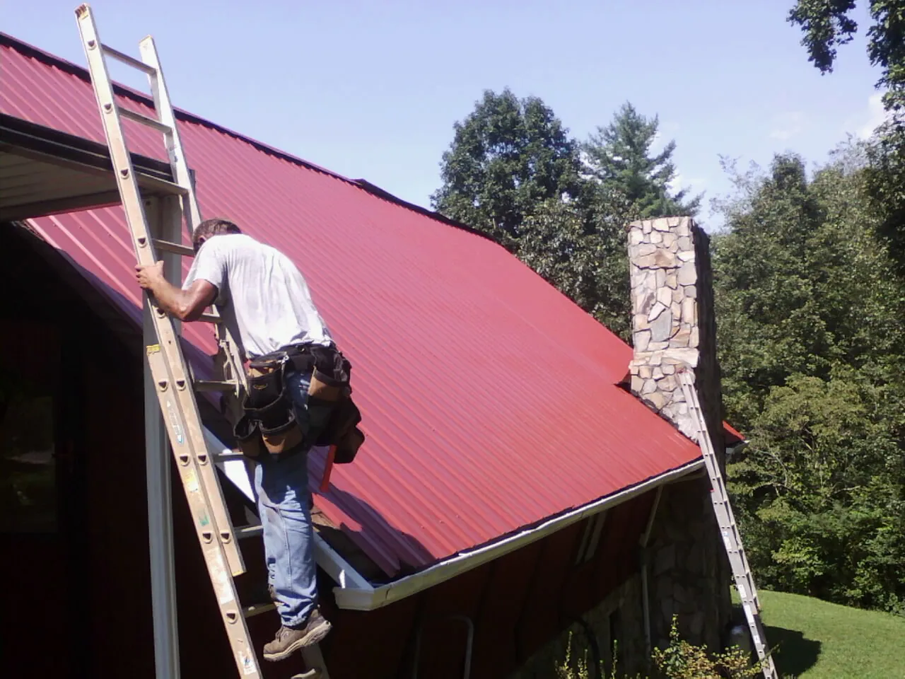 Roofer on a ladder beside a red metal roof and stone chimney — Soffit & Fascia Repair by Roofing Brothers in Audubon